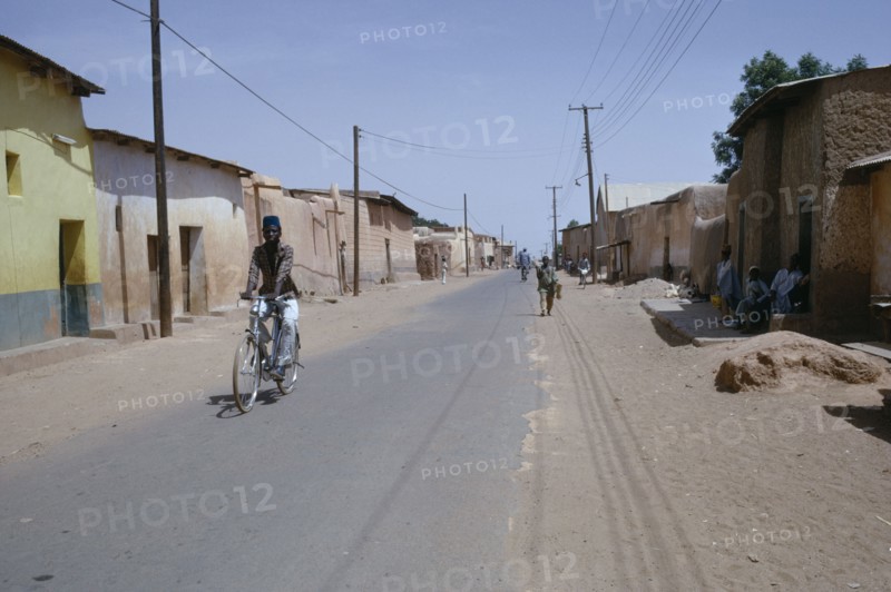 NIGERIA, Kano, Street scene in old city with cyclist on road lined with mud brick architecture.