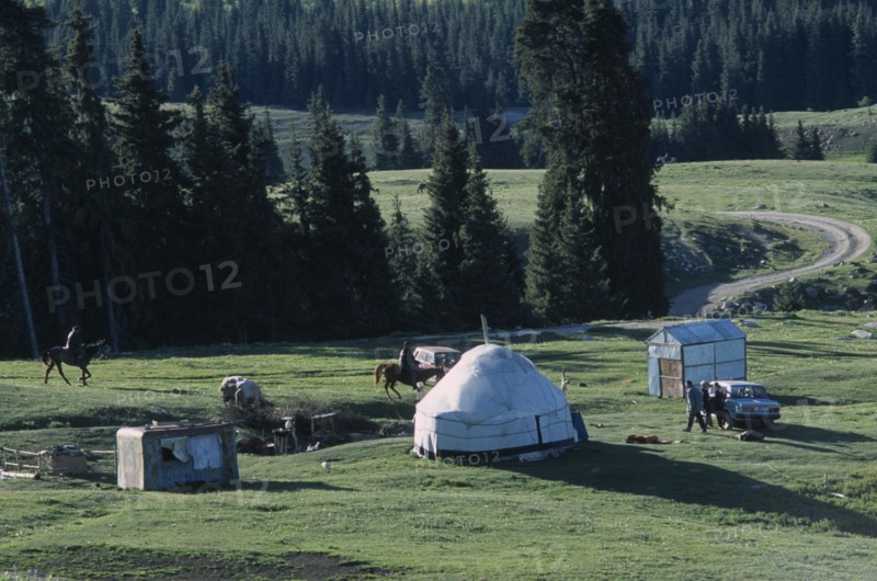 KIRGHIZSTAN, Tyan Shan, "Two men on horses, some standing around a car next to a yurt and small sheds."