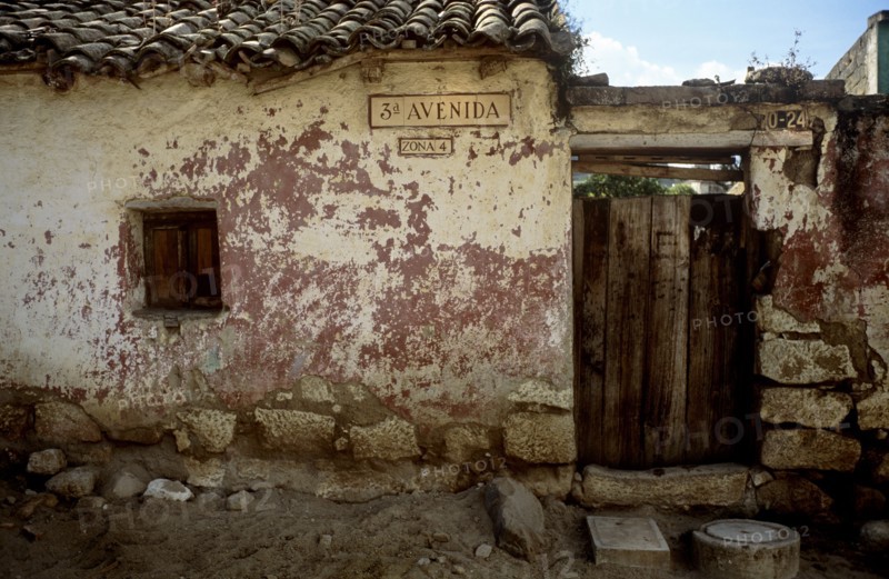 GUATEMALA, Quetzatenango, Xela, "A back street in the outskirts of the Guatemalan town of Xela. A characteristic but crumbling house sits baking in the sun surrounded by large rocks and a dry dusty road. The house itself is worn and crumbling. Some of the tiles on the roof are beginning to crack and the old red brickwork can be seen behind flaking paint. On the front wall of the house hangs the road and area name - '3 Avenida, Zona 4' The entrance to the house consists of an old but firm wooden gate. The gate, made of odd lengths of wood is closed but one can just make out the green foliage in the background. The sky is mainly clear and blue and to the left of the road name sits a small hole in the front of the house with a closed shutter in."