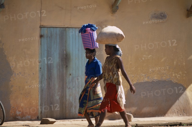 MADAGASCAR, Ambalavao, Two women wearing colourful clothing walking along a street next to a building one carrying a shopping basket on her head and the other woman carrying a bag of grain