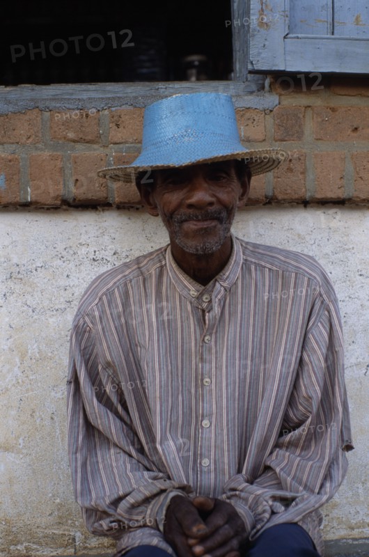 MADAGASCAR, Ihosy, Portrait of an elderly man sitting against a wall wearing a blue straw hat