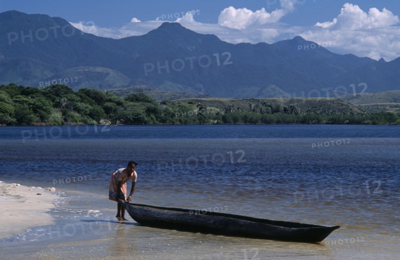 MADAGASCAR, Fort Dauphin, Lokaro, A woman standing at the waters edge of a sandy beach about to push her Pirogue back into the water to return to her village with mountains in the distance