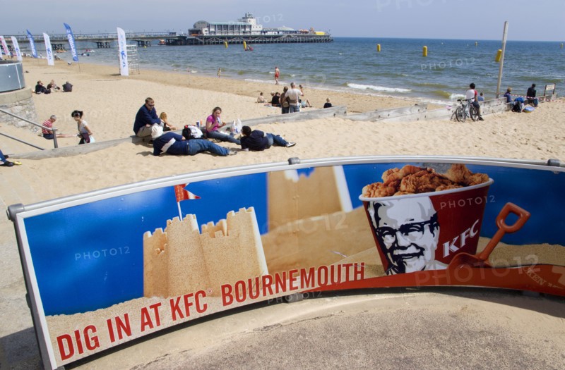 ENGLAND, Dorset, Bournemouth, Tourists playing in the sand and watching speedboat races off the West Beach beside the pier. A KFC advertising board in the foreground.