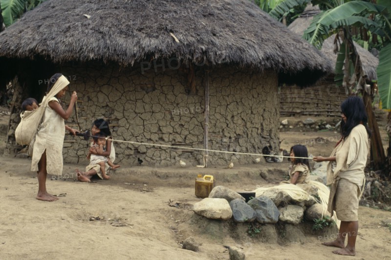 COLOMBIA, Sierra Nevada de Santa Marta, Avinque, Parque Nacional. Kogi-Wiwa family spinning rope from strands of fique cactus fibre. Mother holds spindle and spins whilst father holds out extended new rope. Two young daughters look on whilst baby son sleeps in large woven cotton mochila/shoulder bag on mothers back  his weight taken by broad headband. All family wearing traditional woven cotton mantas/cloaks