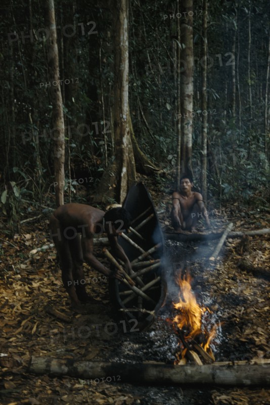 COLOMBIA, Vaupes Region, Tukano Tribe, "2 men making a canoe from a single tree trunk. After initial shaping with axes, canoe burnt and sides expanded in heat with wooden spacers. Burning slims sides, shapes and hardens the wood"