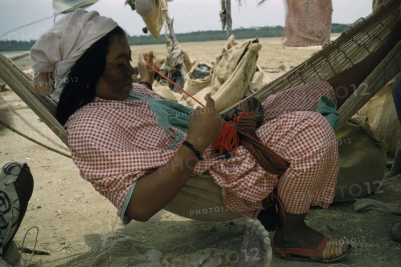 COLOMBIA, Guajira Peninsula, Guajiro / Wayuu Tribe, Wayuu woman sews a coloured “faja” / woven woollen belt for her husband sitting in a fique / cactus fibre hammock in their scanty desert shelter