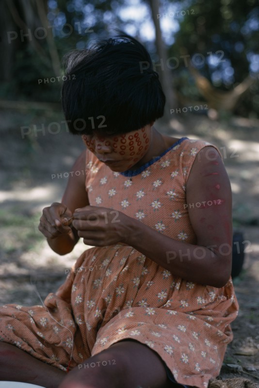 COLOMBIA, Llanos Plains, Rio Meta Arauca, "rio Meta ""Tree Camp"". Cuiva woman  Yanakwa's wife  wearing traded flower print dress with red Achiote facial paint She threads coloured glass beads obtained from boats which ply the rivers of the Llanos  buying local produce and selling trade goods  Llanos plains running East from Andes to Orinoco. rio Meta  Arauca Indigenous Tribes Cuiva  "
