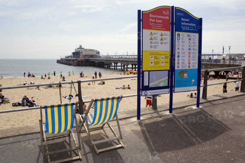 ENGLAND, Dorset, Bournemouth, Deckchairs beside an information sign on The East Beach promenade with Bournemouth Pier beyond tourists on the sandy beach and paddling in the sea.
