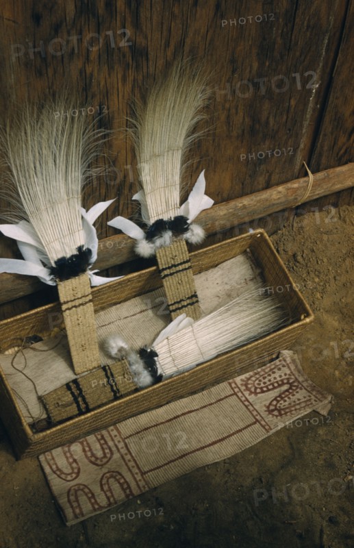 COLOMBIA, Vaupes Region, Tukano Tribe, "Sacred box of ceremonial feather head-dresses, ritual adornments, hung from “maloca” longhouse roof and brought down only for festivals /dances. Royal crane tail feather headdress, dyed bark-cloth apron in foreground"