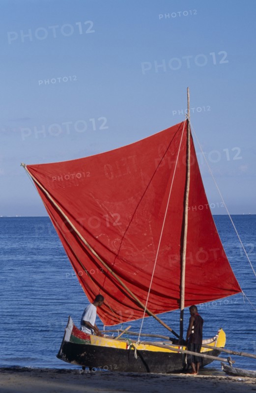MADAGASCAR, Tulear, Ifaty Beach. Men with a red sail Pirogue boat at the waters edge