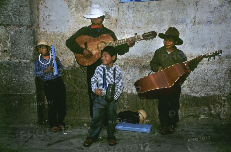 MEXICO, Oaxaca State, Oaxaca, " In a backstreet of the colonial town of Oaxaca stands a local family playing traditional Mexican music to an eclectic group of enthusiastic passer-bys. The father seems to be leading the musical band of four and stands proudly at the back against a grey brick wall. He sports a white cowboy hat and blows on a harpsichord whilst strumming his acoustic guitar. On his right hand side stands his oldest son, dressed in similar attire strumming a much larger guitar that looks more like a cello. On the left of shot, there is the middle son who unenthusiastically leans in the corner, again dressed in a beige cowboy hat, a checkered shirt and dark torusers. He is playing a plastic wind instrument which also appears to have some kind of keyboard. In front of these three stands the youngest of the group. The young child wears a checkered blue and white shirt and seems to be shputing and gesturing at the passing trade for tips or 'propinas'. The ground below them lie...