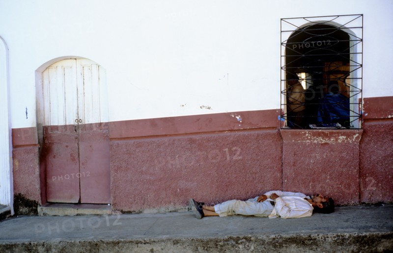 EL SALVADOR, Suchitoto, "In the heart of this old colonial town lies a colourful backstreet. The outside walls of the houses slopes down from right to left and at the front of shot one can make out the edge of a bumpy pavement. The wall in the background has been painted halfway in a rustic red colour. The wooden doorway is visibly closed and there are iron bars in front of the only visable window. Behind the bars is a local barbers as one can just about make out the shapes of a barber and his client sitting down. On the pavement on the foreground lies a local dressed from head to toe in beige linen. He is sleeping on the dusty ground with his legs crossed and his hands lying gently on his chest. His skin is dark and worn and although his position seems a little unorthodox, on the whole he seems surprisingly comfortable. "