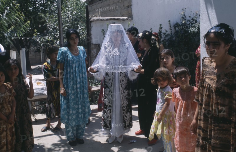 TAJIKISTAN, Wedding, "The bride in a white dress, veil and decorated jacket surrounded by guests at a village wedding."