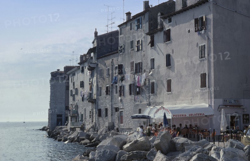 CROATIA, Istria, Rovinj, Old city waterfront building with a washing line hanging from shuttered windows and people sitting outside under the sun awning of a Pizzeria restaurant attached to the bottom of the building. Large sea defence boulders lining the waters edge