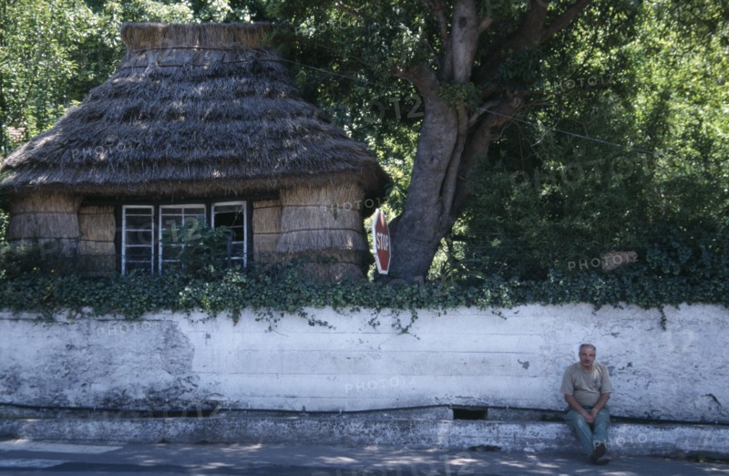 PORTUGAL, Madeira, Camacha, Traditional thatched cottage beside the Largo do Achada the central square with a man sat on the pavement next to a white wall
