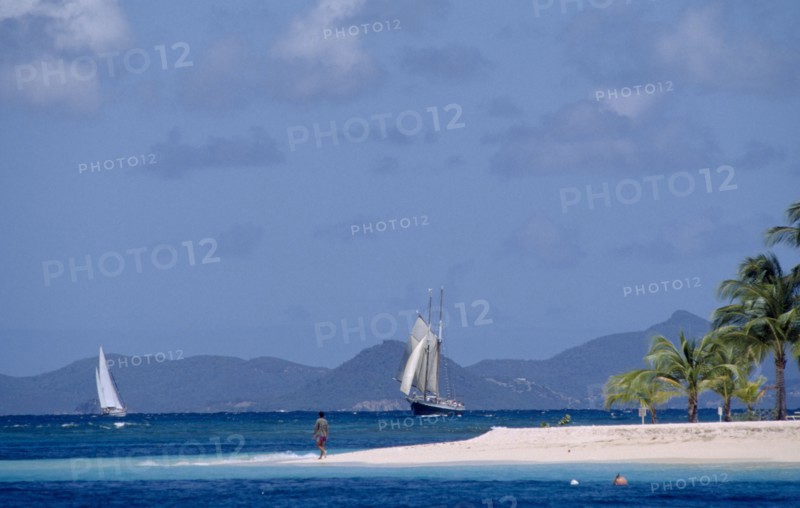 GRENADINES, Petit St Vincent, Palm Island Resort. Man walking on sandy outstretch of beach surrounded by turquoise sea with boats on water