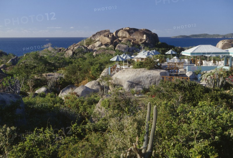 BRITISH VIRGIN ISLANDS, Virgin Gorda, The Baths. Granite boulders amongst plants and vegetation with chairs and sun shades from a restaurants open air verandah in the middle. The ocean seen in the distance