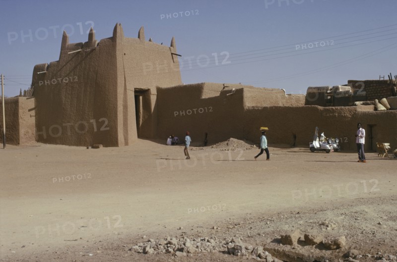 NIGERIA, Kano, Ancient mudbrick city walls and passing people and street trader carrying tray of fruit on his head.