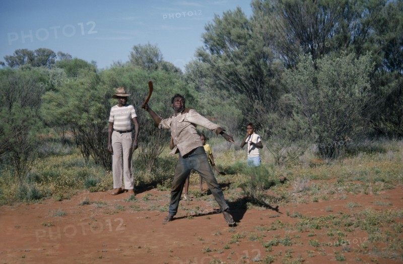 AUSTRALIA, Northern Territory, Alice Springs, Aborigine man throwing a Boomerang with a man and children standing behind him looking on
