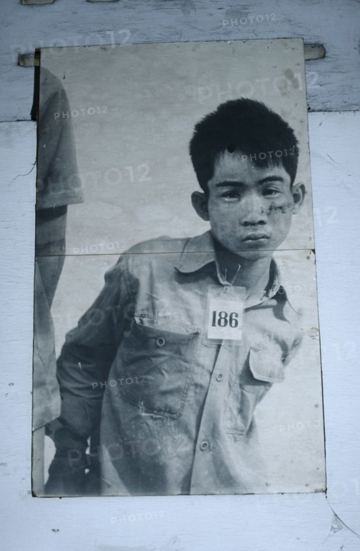 CAMBODIA, Phnom Penh, Tuol Sleng Museum. Photograph of a young male prisoner in a Khmer Rouge prison wearing a number tag around his neck