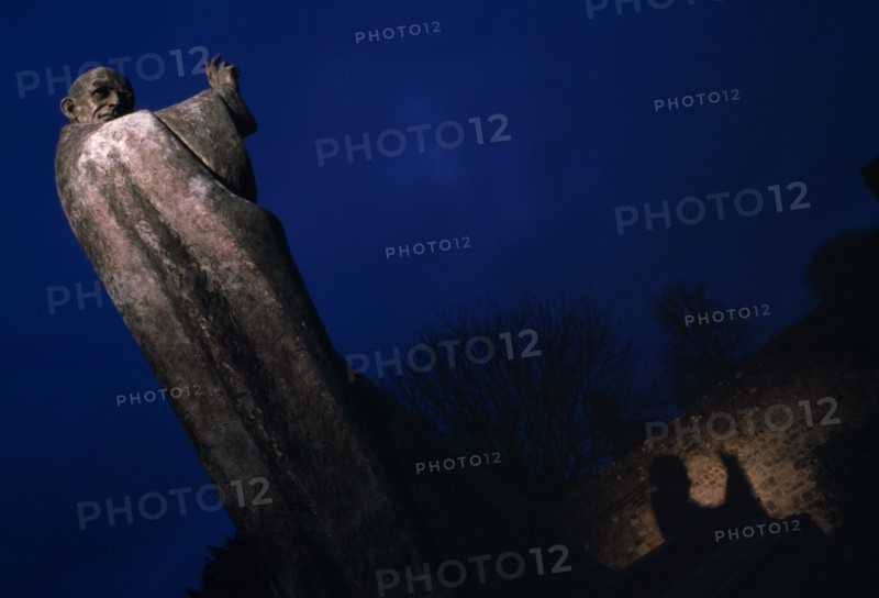 ENGLAND, West Sussex, Chichester, Statue of Saint Richard partially illuminated at night in Cathedral grounds with shadow reflecting on nearby wall.
