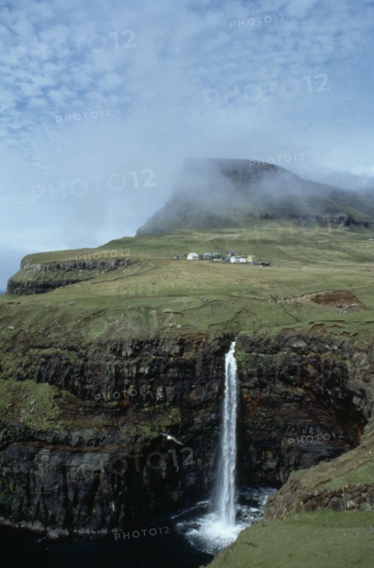 DENMARK, Faeroe Islands, River Gazadalla, Waterfall and rocky coastline with houses built on green landscape