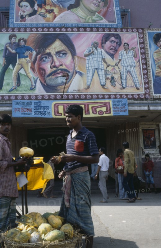 BANGLADESH, Dhaka, Cinema with colourful poster hoadings above entrance and men at a fruit stall in the foreground