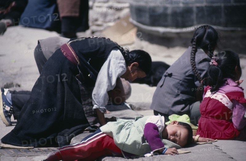 TIBET, Lhasa, Pilgrims at Jokhang Monastery.  Kneeling woman and prostrate child on ground in front of temple entrance.