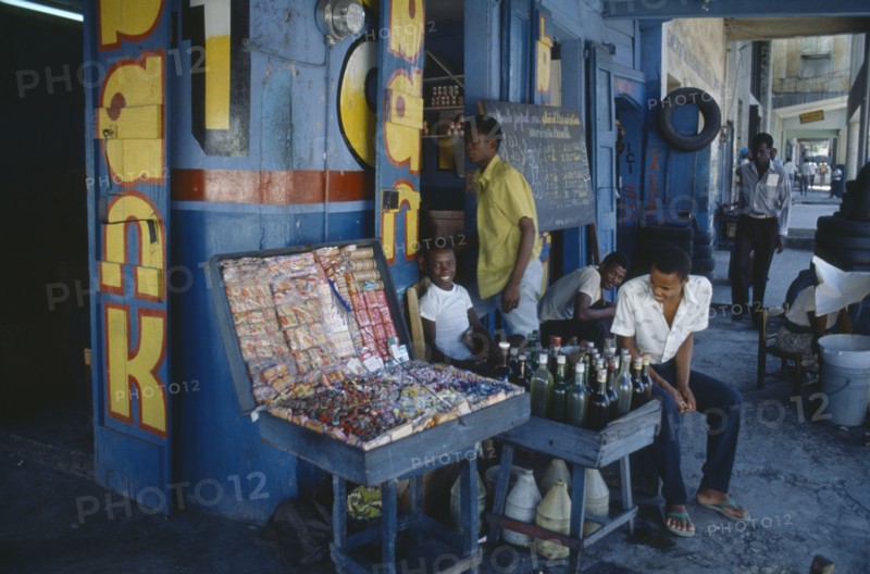 HAITI, Port au Prince, Children and young men outside shop of sweets and drinks vendor with blue and yellow painted exterior.