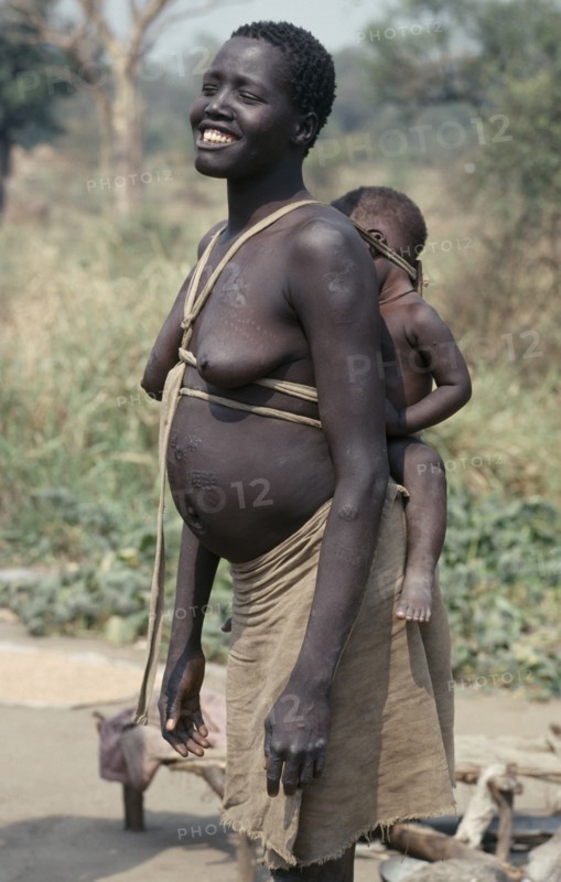ETHIOPIA, West, Gambella, Uduk refugees from Sudan.  Smiling pregnant woman carrying baby on her back in cloth and wood sling.  Note scarification on stomach.