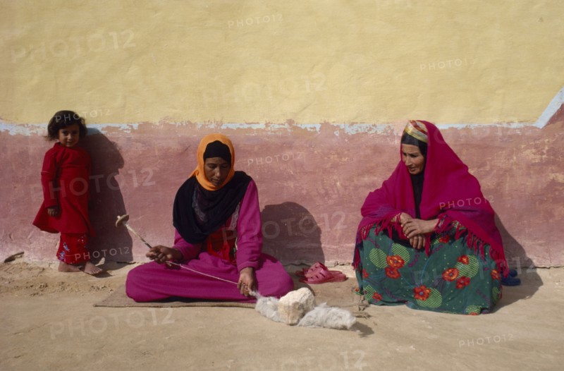 EGYPT, Western Desert, Bedouin, "Women and child in colourful dress spinning wool, sitting against faded yellow and ochre coloured wall of building."