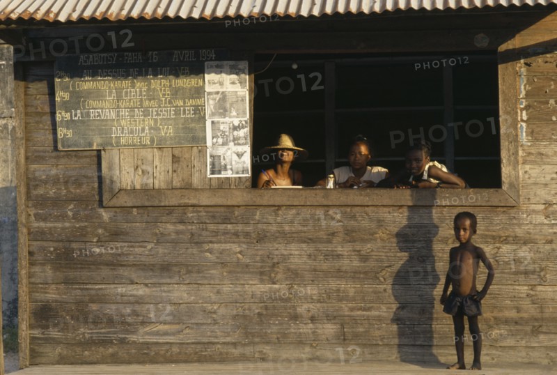 MADAGASCAR, Fort Dauphin, Child standing outside local cinema ticket office with three girls at open window and blackboard on outside wall with film listings in French.