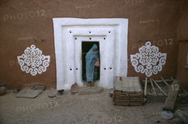 MAURITANIA, Oualata, "Woman stepping out from doorway with white surround and traditional bas relief motifs on each side of applied gypsum, white and red clay."