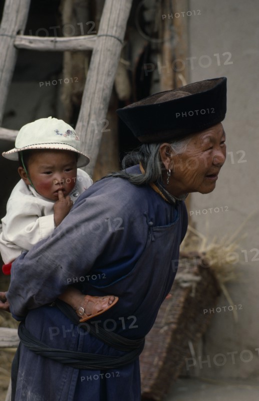 CHINA, Qinghai Province, Huzhu District, Elderly Tu nationality Lamaist woman of Yellow Hat Buddist sect carrying child on her back.