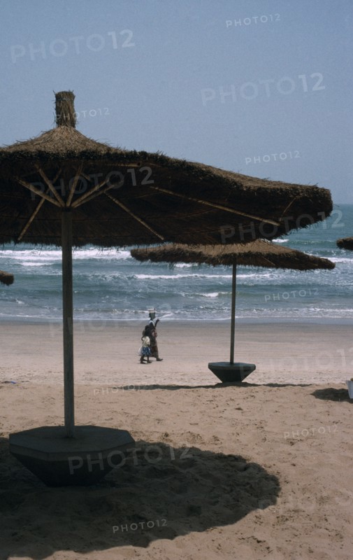 GAMBIA, Landscape , Beach, Beach with straw sun shades and a woman with children walking along the sand
