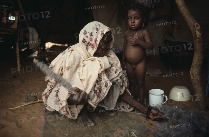 SUDAN, North Kordofan, Work, Kababish nomad woman spinning wool inside hut with little girl standing beside her.