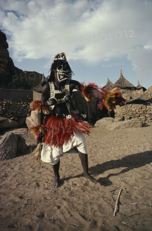 MALI, Ceremony, "Portrait of Dogon dancer wearing costume of short fringed red and yellow skirt, false breasts and mask decorated with cowrie shells."