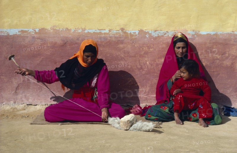 EGYPT, Western Desert, Settled Bedouin woman spinning wool with older woman and child sitting beside her against faded ochre and terracotta painted wall.