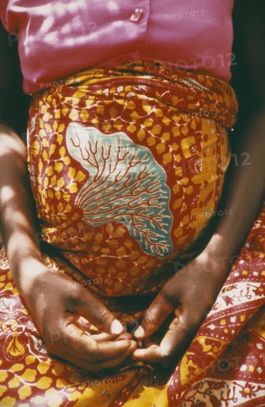 GHANA, Volta Region, Cropped shot of woman thirty six weeks pregnant wearing brightly coloured sarong and with her hands folded in her lap beneath her belly.