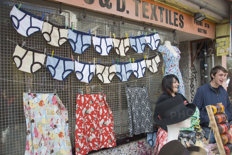 ENGLAND, East Sussex, Brighton, A North Lanes market stall selling hats and clothes with a  large colourful display of underwear behind two young smiling stall holders.