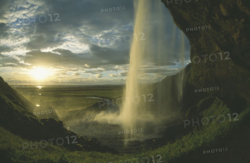 ICELAND, Seljalandfoss, View from behind waterfall in low golden sunlight through drifting cloud.