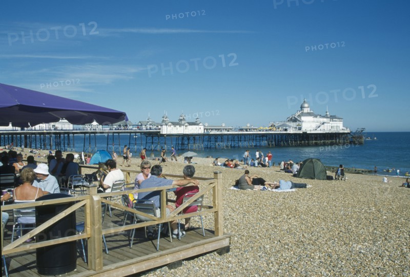 ENGLAND, East Sussex, Eastbourne, Beach cafe with people sat on chairs under parasols infront of busy shingle beach and pier