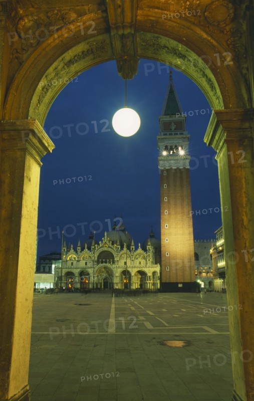ITALY, Veneto, Venice, Piazza di San Marco.  Part view of square with Basilica di San Marco and the Campanile framed by stone archway with central circular lamp illuminated at night.