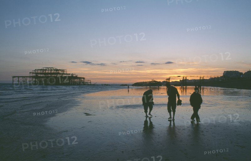 ENGLAND, East Sussex, Brighton, The ruined West Pier at sunset with low tide and a group of people walking together along beach.