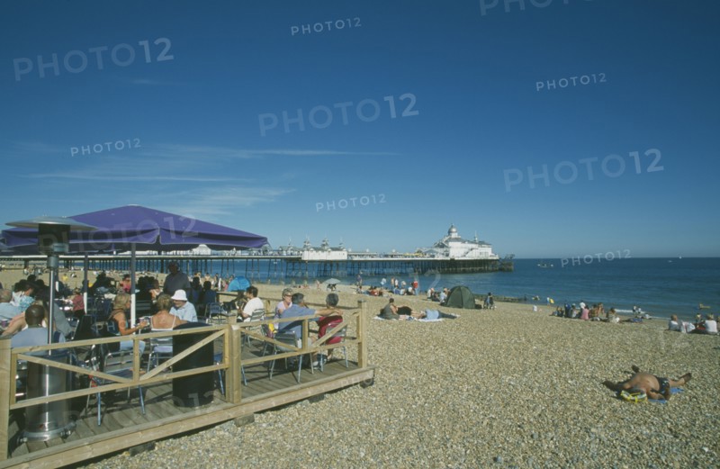 ENGLAND, East Sussex, Eastbourne, Beach Cafe with people sat on chairs under parasols and the pier in the distance.