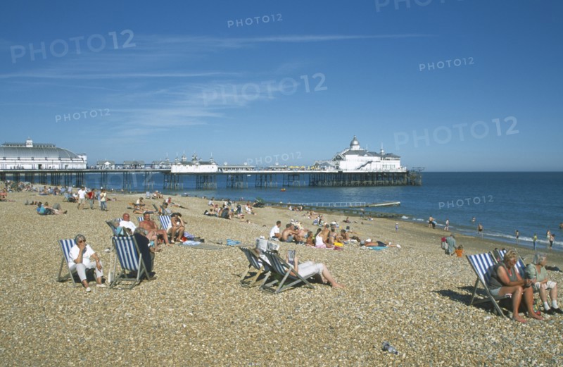 ENGLAND, East Sussex, Eastbourne, View across shingle beach with people sat on deckchairs towards the pier.