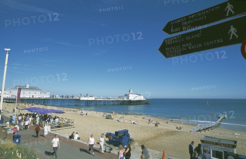ENGLAND, East Sussex, Eastbourne, View across busy seafront towards shingle beach and pier with direction signs for tourist attractions in the forground.