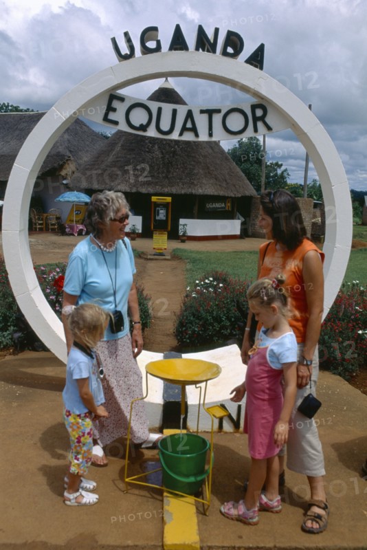 UGANDA, Equator, European family watching water go down plughole beside sign.