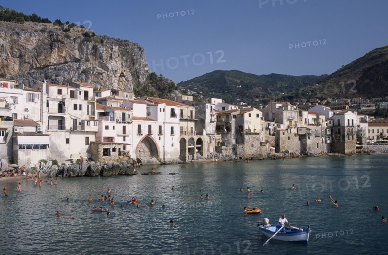 ITALY, Sicily, Palermo, Cefalu. View across people swimming in the sea next to sandy beach with a man rowing a small boat in the foreground overlooked by waterfront apartments and hills behind