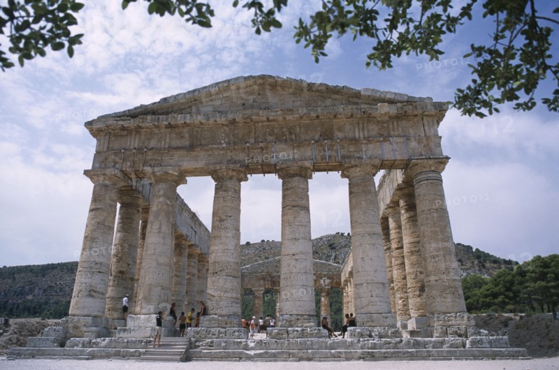 ITALY, Sicily, Segesta, Doric Temple. Ruins of the ancient city. Colonnaded structure framed by tree branches with visitors walking around